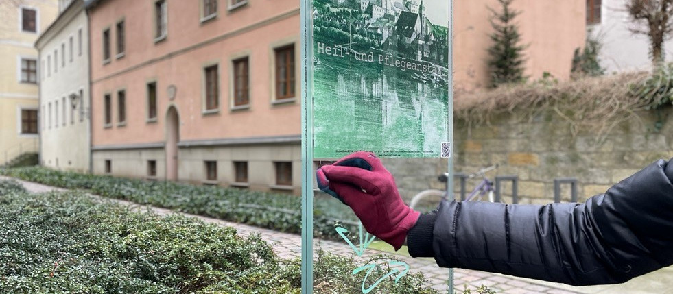 KI generiert: Das Bild zeigt eine Person in schwarzer Jacke mit roten Handschuhen, die mit einem pinken Stift auf eine Glasfläche schreibt oder zeichnet. Im Hintergrund sind ein Gebäude und ein Garten mit Büschen zu sehen.