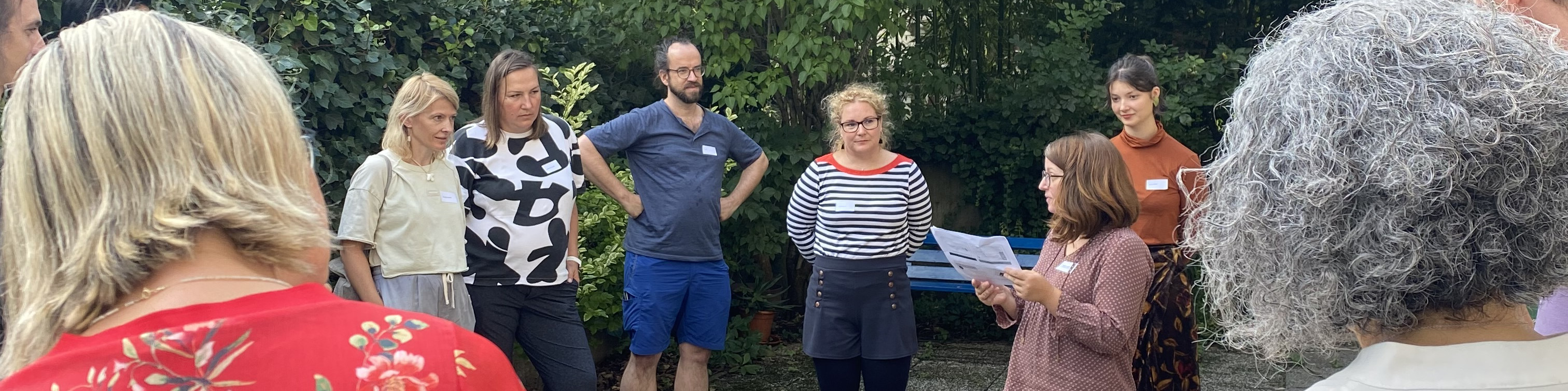 KI generiert: Das Bild zeigt eine Gruppe von Personen im Freien, die sich um eine Frau versammelt haben, die ein Blatt Papier hält und anscheinend etwas vorliest oder erklärt. Der Hauptinhalt des Bildes ist eine informelle Zusammenkunft oder Diskussion in einem Garten oder Park.