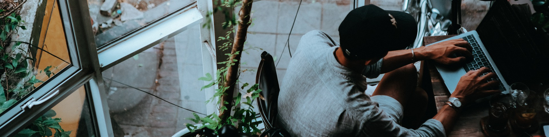 KI generiert: Das Bild zeigt eine Person, die an einem Laptop arbeitet, während sie an einem Tisch in der Nähe eines großen Fensters sitzt. Der Hauptinhalt des Bildes ist die Arbeitsumgebung mit Fokus auf die Person und ihren Laptop.