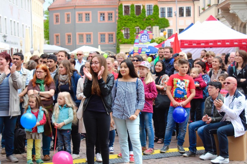 KI generiert: Das Bild zeigt eine Menschenmenge bei einem öffentlichen Event, bei dem zwei junge Frauen im Vordergrund stehen und sich unterhalten. Im Hintergrund sind bunte Zelte und historische Gebäude zu sehen, während einige Kinder mit Ballons spielen.