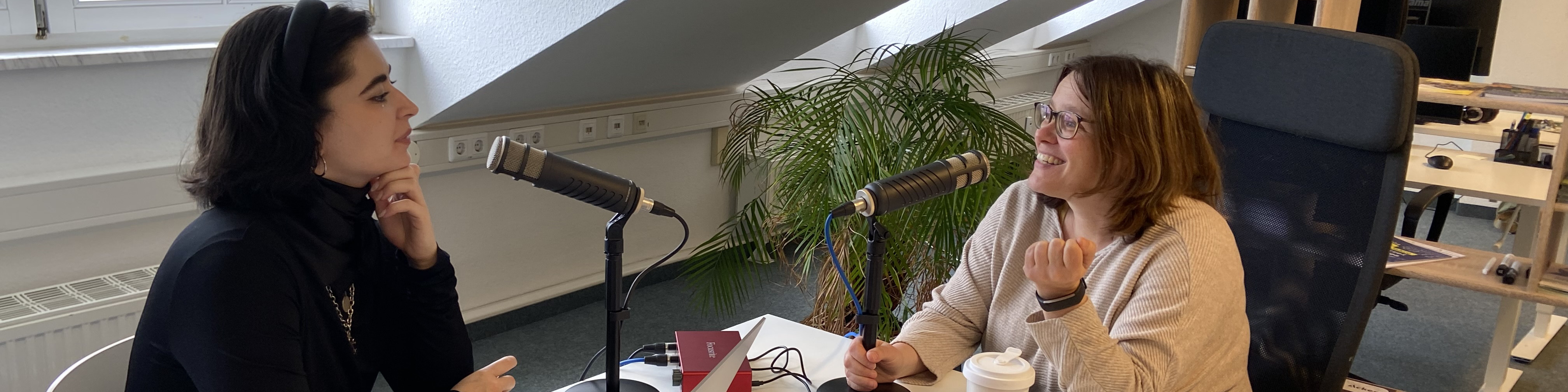 KI generiert: Das Bild zeigt zwei Frauen, die sich in einem Büro oder Studio unterhalten, wobei eine Frau an einem Tisch mit Mikrofonen sitzt und die andere Frau ihr gegenüber steht. Der Hauptinhalt des Bildes ist ein Gespräch oder Interview, das möglicherweise für eine Podcast- oder Radioaufnahme stattfindet.