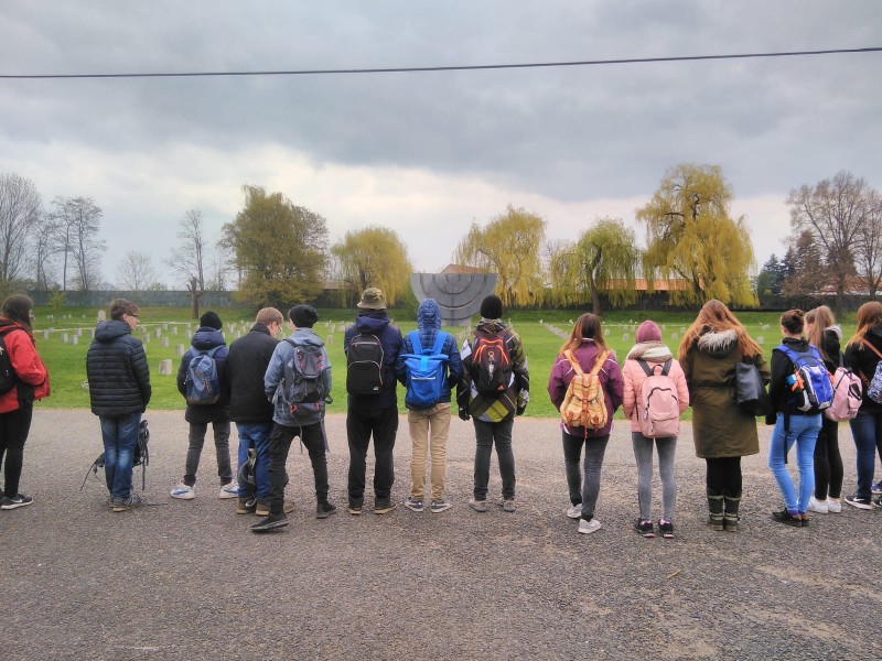 KI generiert: Das Bild zeigt eine Gruppe von Personen, die auf einem Weg stehen und in Richtung eines Denkmals oder einer Skulptur in einer parkähnlichen Umgebung mit Bäumen und Grabsteinen blicken. Der Hauptinhalt des Bildes ist die Gruppe von Menschen, die sich anscheinend eine historische oder kulturelle Stätte anschauen.