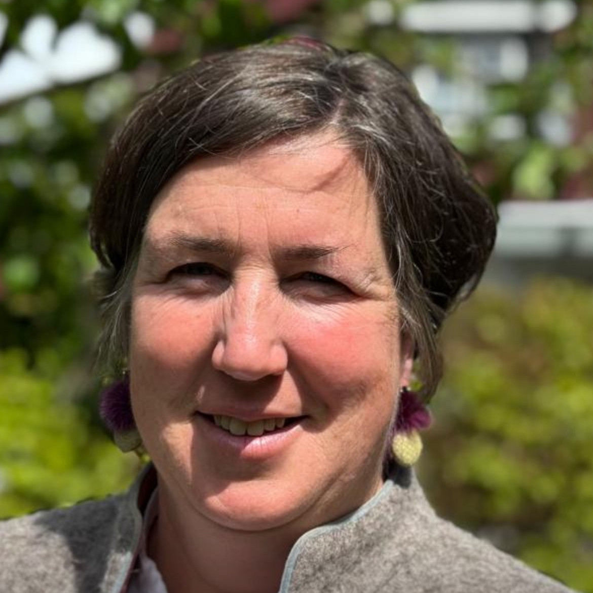 KI generiert: Das Bild zeigt eine lächelnde Frau mit kurzen, dunklen Haaren und grauer Jacke, die sich in einer grünen, natürlichen Umgebung befindet. Der Hauptinhalt des Bildes ist das Porträt der Frau im Freien.