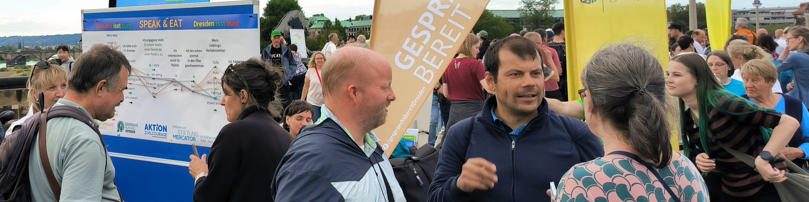 KI generiert: Das Bild zeigt eine Gruppe von Menschen, die sich auf einer Veranstaltung unterhalten, mit einem Banner, das "GESPRÄCHE BEREIT" anzeigt. Im Hintergrund sind weitere Personen und ein Plakat mit dem Titel "SPEAK & EAT" zu sehen.