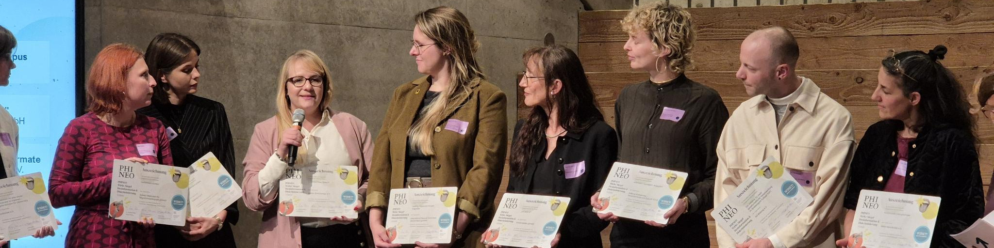 KI generiert: Das Bild zeigt eine Gruppe von Personen, die stolz ihre Zertifikate präsentieren, während eine Frau in der Mitte ein Mikrofon hält und möglicherweise eine Rede hält. Der Hauptinhalt des Bildes ist eine Zertifikatsübergabezeremonie.