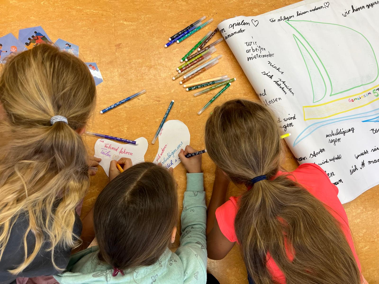 KI generiert: Das Bild zeigt drei Kinder, die an einem Tisch sitzen und gemeinsam an einem Projekt arbeiten. Sie malen und schreiben auf Papier, während verschiedene Stifte und Schulmaterialien auf dem Tisch verstreut liegen.
