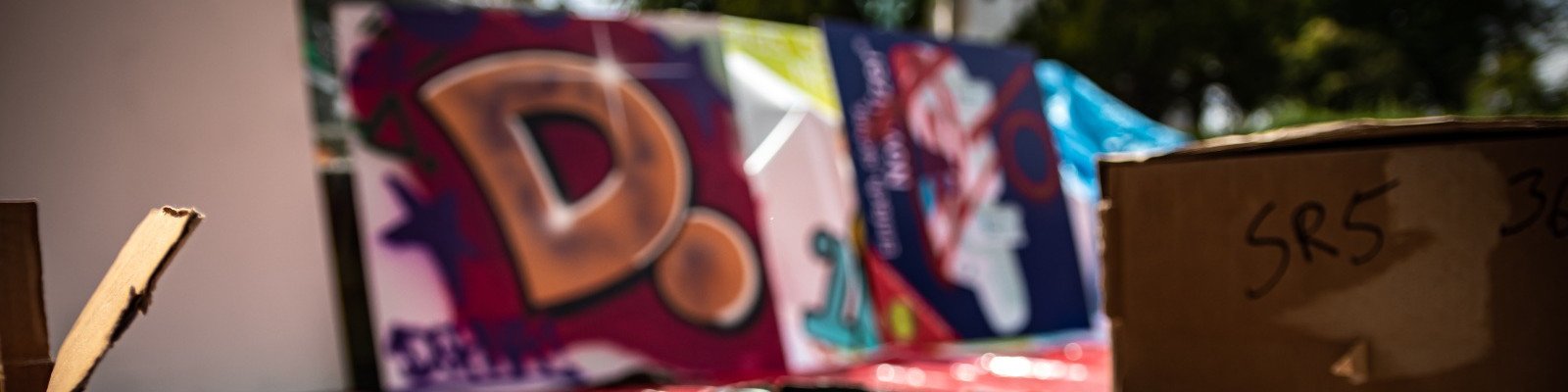 KI generiert: Das Bild zeigt einen unscharf fotografierten Ausschnitt eines bunten Graffiti mit einem großen, stilisierten Buchstaben "D". Im Vordergrund sind einige Pappkartons zu sehen, auf denen teilweise Schriftzeichen zu erkennen sind.