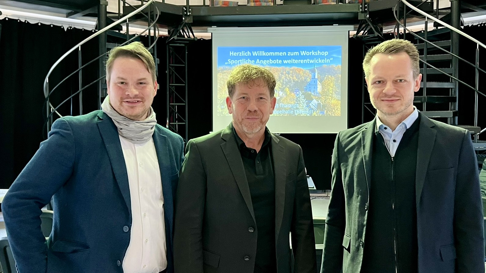 KI generiert: Das Bild zeigt drei Männer in Businesskleidung, die vor einem Projektionsbildschirm mit der Aufschrift "Herzlich Willkommen zum Workshop 'Sportliche Angebote weiterentwickeln'" stehen. Der Hauptinhalt des Bildes ist die Präsentation oder der Workshop zu sportlichen Angeboten.