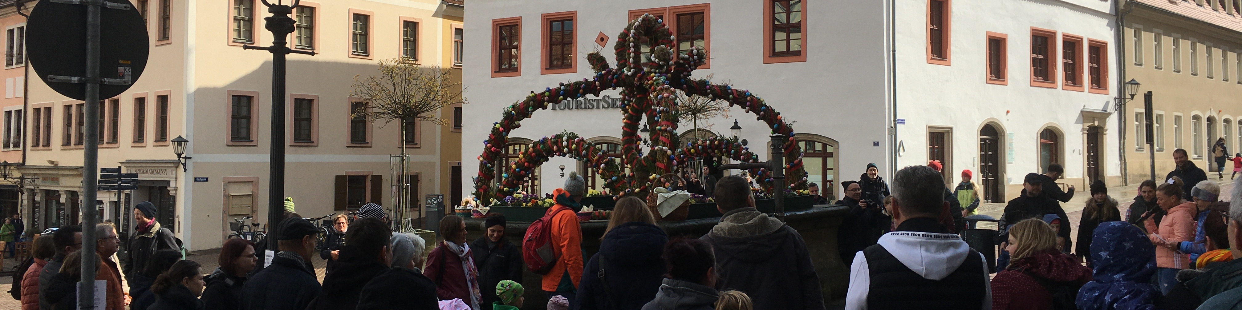 KI generiert: Das Bild zeigt eine belebte Straßenparade mit vielen Menschen, die sich um einen geschmückten Wagen mit einem großen, verzierten Kranz versammelt haben. Im Hintergrund sind historische Gebäude zu sehen, was auf eine traditionelle Feier oder einen Festumzug in einer Altstadt hindeutet.