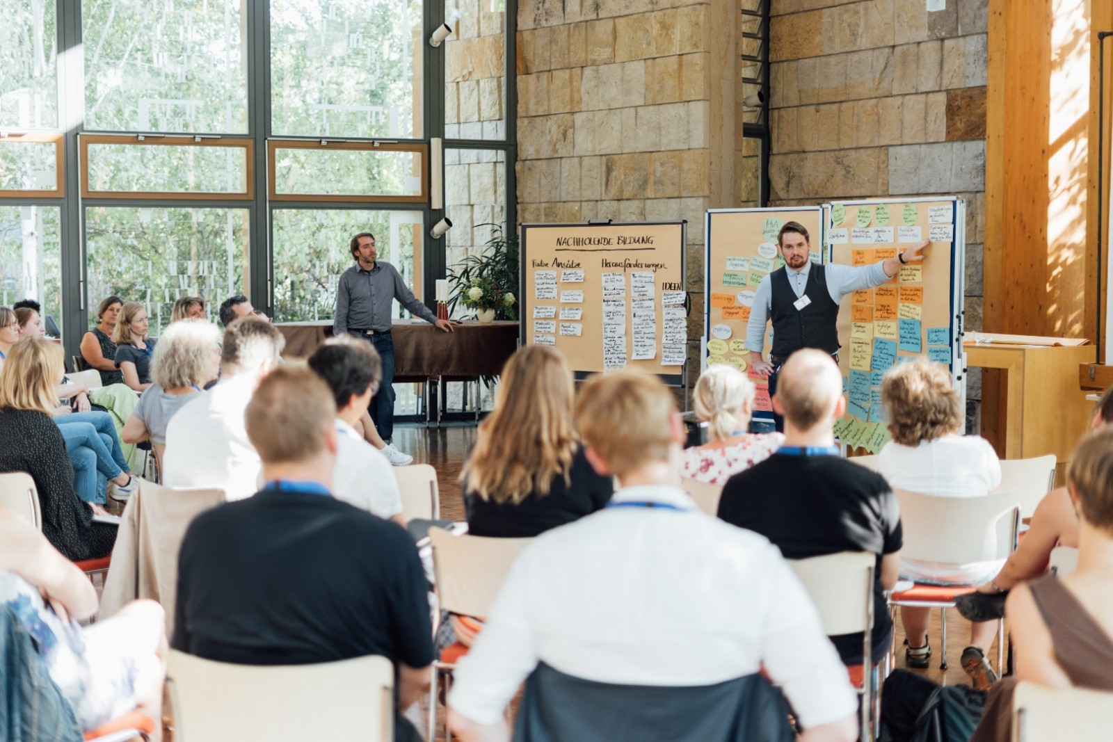 KI generiert: Das Bild zeigt eine Gruppe von Menschen in einem Seminarraum, wobei zwei Männer vorne stehen und eine Präsentation halten. Der Hauptinhalt des Bildes ist eine Schulung oder ein Workshop, bei dem die Teilnehmer aufmerksam zuhören und Notizen machen.