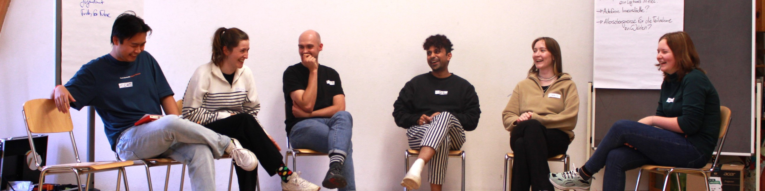 KI generiert: Das Bild zeigt eine Gruppe von sechs Personen, die auf Stühlen in einer Reihe sitzen und sich in einer lockeren Runde unterhalten. Der Hauptinhalt des Bildes ist eine informelle Diskussion oder ein Workshop in einem Innenraum.