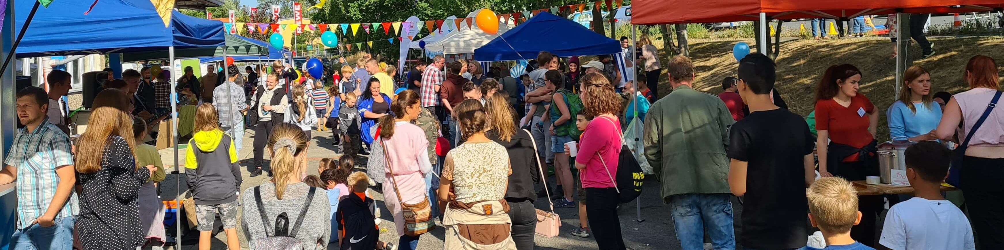 KI generiert: Das Bild zeigt eine belebte Outdoor-Veranstaltung mit vielen Menschen, die zwischen bunten Zelten und Ständen umherlaufen. Der Hauptinhalt ist ein Fest oder Markt mit einer lebhaften und fröhlichen Atmosphäre.
