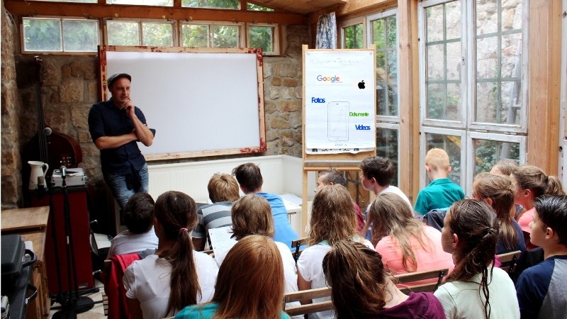KI generiert: Das Bild zeigt einen Mann, der vor einer Gruppe von Kindern und Jugendlichen steht und eine Präsentation hält. Im Hintergrund sind ein Whiteboard und eine Pinnwand mit Logos von Unternehmen wie Google, Apple, Folios, Edumoto und Velux zu sehen.