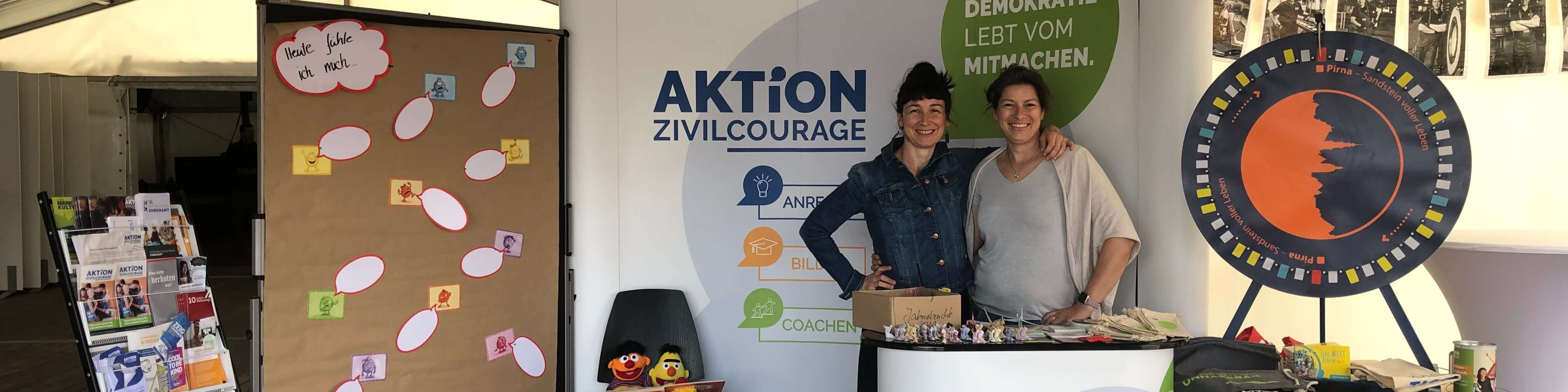 KI generiert: Das Bild zeigt einen Stand der "Aktion Zivilcourage" auf einer Veranstaltung, an dem zwei Personen stehen und Informationsmaterial anbieten. Im Hintergrund sind ein Plakat mit der Aufschrift "Demokratie lebt vom Mitmachen" und ein Glücksrad zu sehen.
