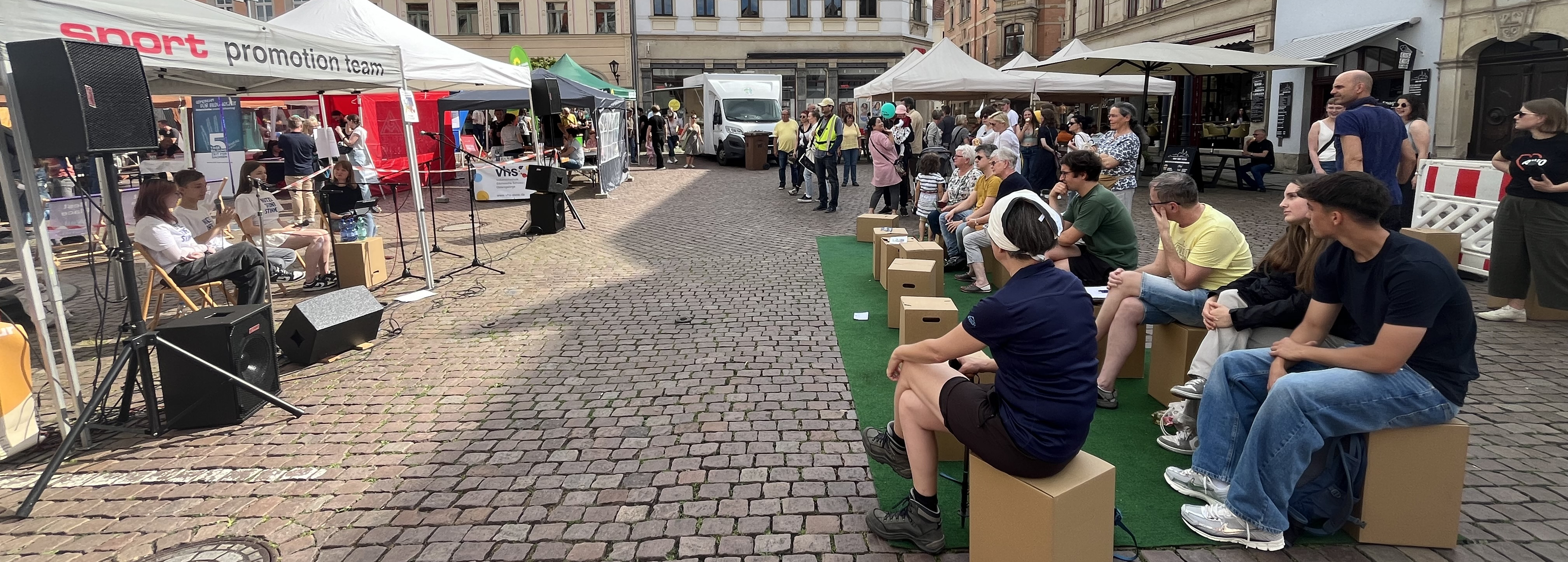 KI generiert: Das Bild zeigt eine lebendige Straßenveranstaltung mit Menschen, die auf Holzbänken sitzen und Musik hören, während im Hintergrund weitere Besucher und Stände zu sehen sind. Der Hauptinhalt des Bildes ist eine öffentliche, gesellige Zusammenkunft mit Musik und verschiedenen Aktivitäten.