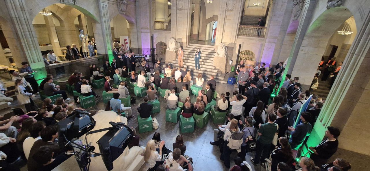 KI generiert: Das Bild zeigt eine große Menschenmenge in einem historischen Gebäude mit Säulen und einer Treppe, die an einer Veranstaltung teilnimmt. Im Vordergrund sitzen viele Personen auf grünen Sitzkissen, während im Hintergrund eine Bühne mit Moderatoren und Gästen zu sehen ist.