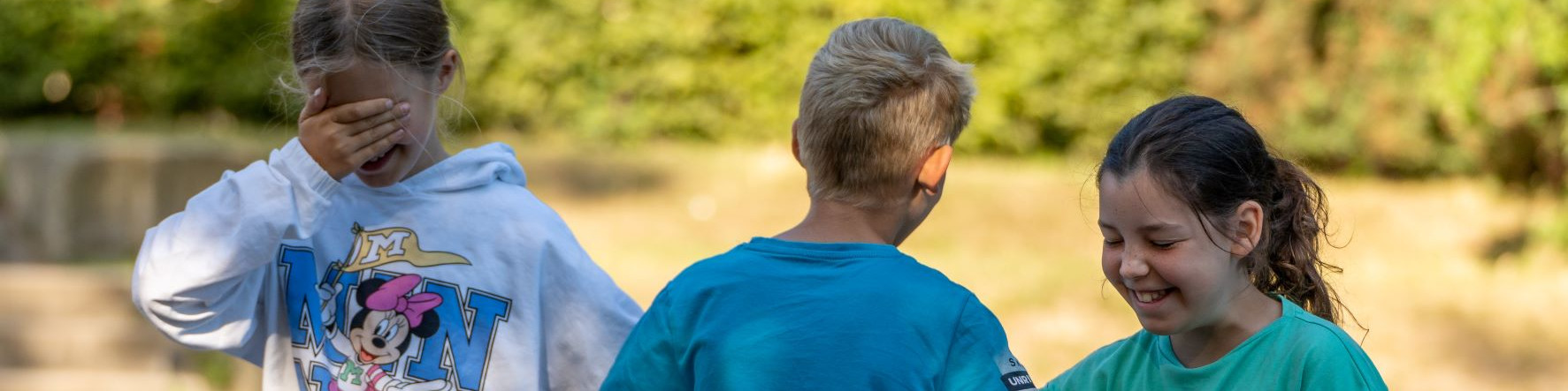 KI generiert: Das Bild zeigt drei Kinder im Freien, die sich unterhalten oder interagieren, wobei eines der Kinder lachend in die Kamera schaut. Der Hauptinhalt des Bildes ist die fröhliche und spielerische Atmosphäre zwischen den Kindern in einer natürlichen Umgebung.