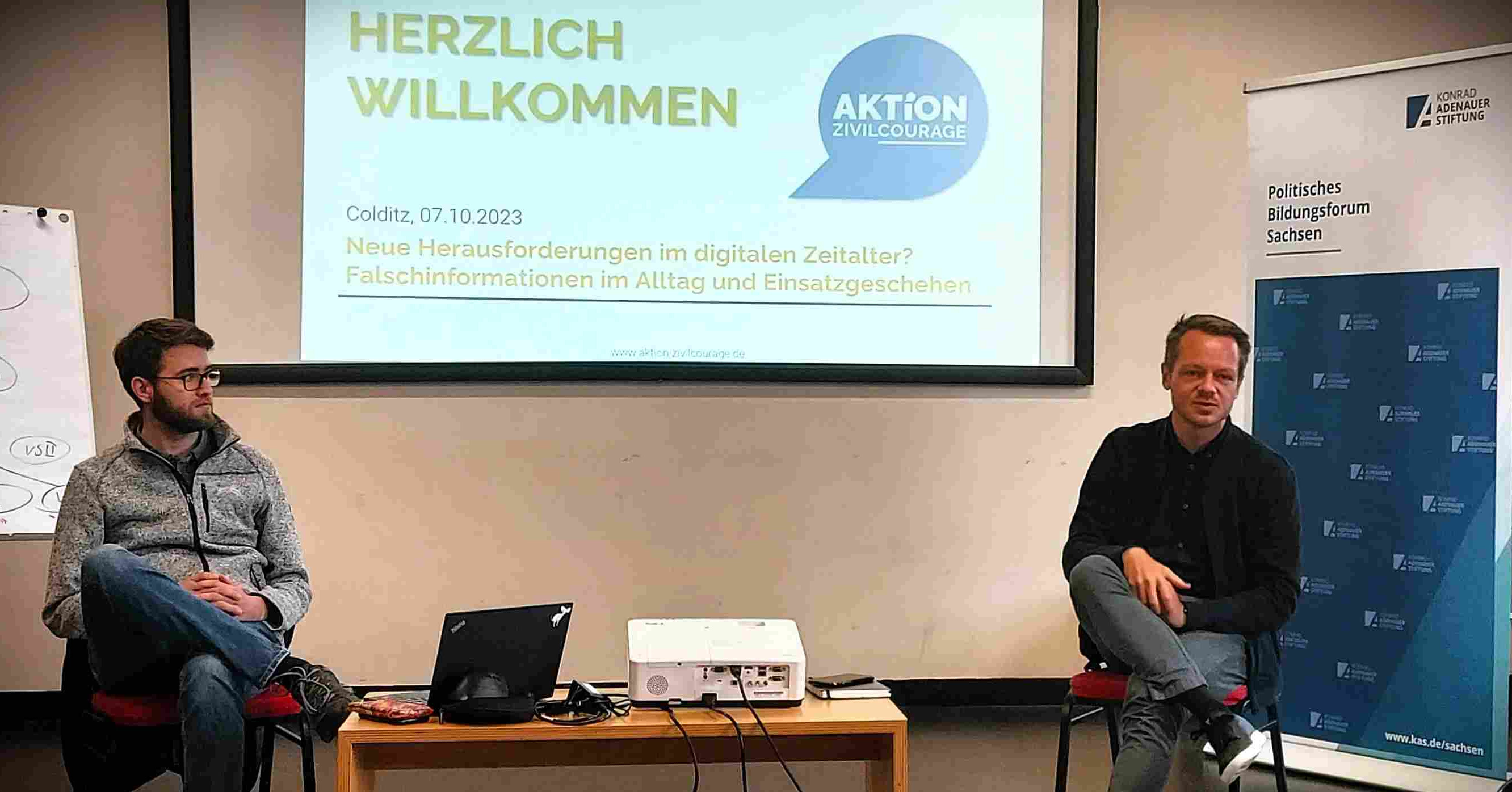 KI generiert: Das Bild zeigt zwei Männer, die auf Stühlen sitzen und sich in einem Raum mit einer Projektion im Hintergrund befinden. Die Projektion enthält den Titel "Herzlich Willkommen" und Informationen über eine Veranstaltung der "Aktion Civilcourage" zum Thema "Neue Herausforderungen im digitalen Zeitalter? Falschinformationen im Alltag und Einsatzgeschehen".