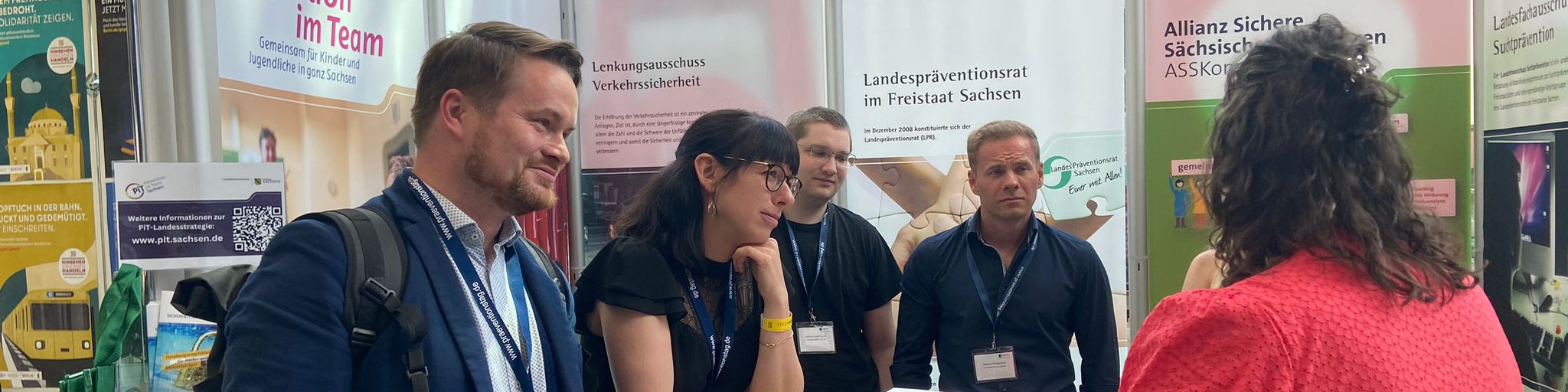 KI generiert: Das Bild zeigt eine Gruppe von Personen auf einer Messe oder Ausstellung, die sich an einem Stand unterhalten. Der Stand hat mehrere Poster mit Informationen, darunter Themen wie Verkehrssicherheit und Präventionsrat im Freistaat Sachsen.