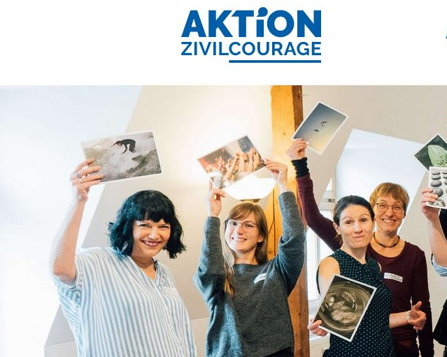 KI generiert: Das Bild zeigt eine Gruppe von Menschen, die fröhlich in die Kamera lächeln und Fotos in die Luft halten. Der Hauptinhalt des Bildes ist eine Gruppe von Personen, die gemeinsam eine positive und enthusiastische Stimmung teilen, möglicherweise bei einem Workshop oder einer Veranstaltung.