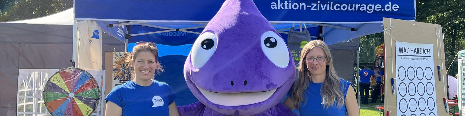 KI generiert: Das Bild zeigt zwei Personen in blauen T-Shirts, die neben einem großen lila Maskottchen in Form eines Tropfens stehen, vor einem Stand mit der Aufschrift "aktion-zivilcourage.de". Im Hintergrund sind ein Glücksrad und ein Plakat mit der Aufschrift "WAS HABE ICH?" zu sehen.