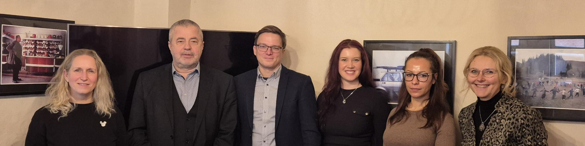KI generiert: Das Bild zeigt eine Gruppe von sechs Personen, die nebeneinander in einem Innenraum stehen und in die Kamera lächeln. Im Hintergrund sind zwei Bildschirme oder gerahmte Bilder an der Wand zu sehen.