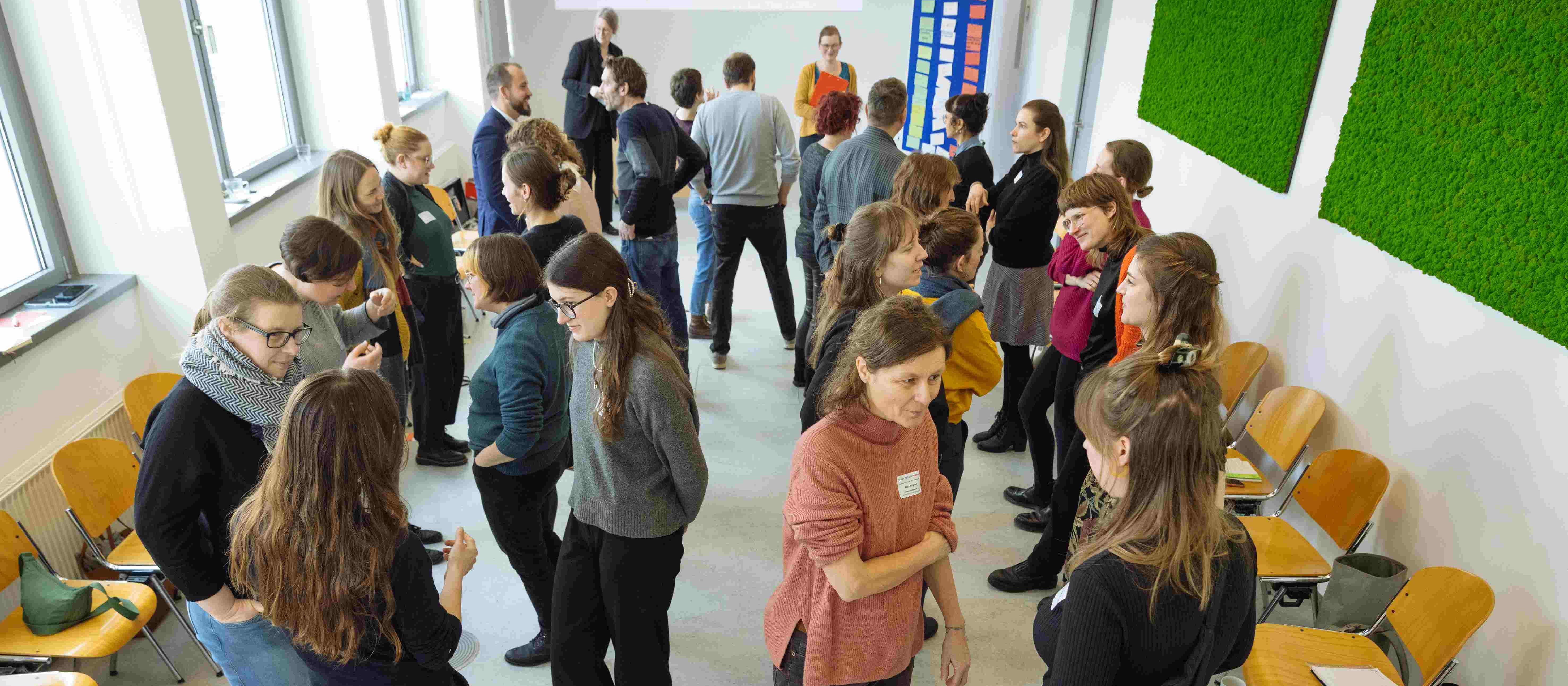 KI generiert: Das Bild zeigt eine Gruppe von Personen, die in einem hellen Raum mit großen Fenstern und grünen Wänden stehen und sich unterhalten. Der Hauptinhalt des Bildes ist eine informelle Zusammenkunft oder ein Netzwerktreffen.
