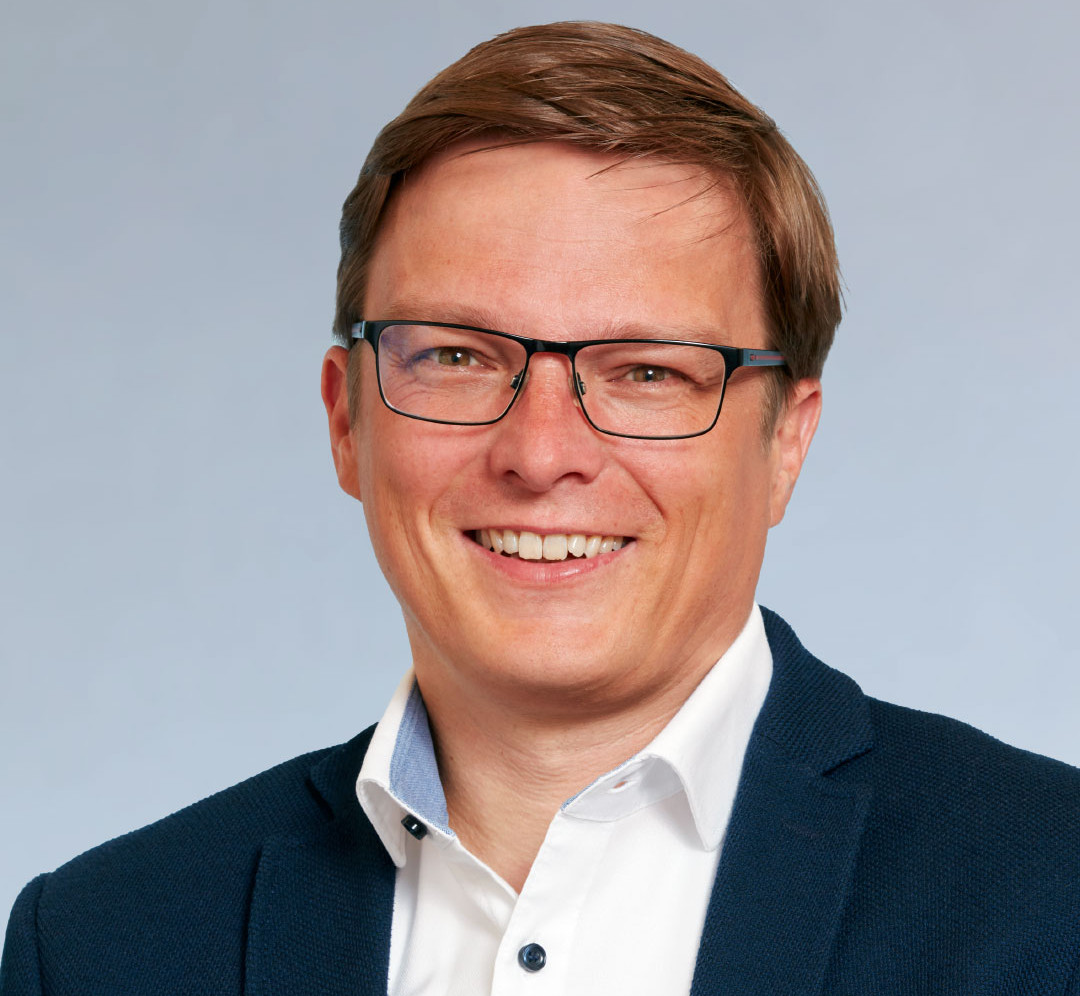 KI generiert: Das Bild zeigt einen lächelnden Mann mit kurzen braunen Haaren und einer Brille. Er trägt ein weißes Hemd mit einem dunklen Blazer und einem blauen Kragen.