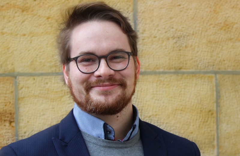 KI generiert: Das Bild zeigt einen lächelnden Mann mit Brille und Bart, der vor einer gelblichen Steinwand steht. Er trägt ein Hemd, einen Pullover und eine Jacke.