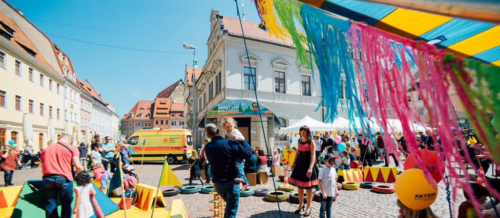 KI generiert: Das Bild zeigt eine lebhafte Straßenveranstaltung in einer historischen Stadt mit bunten Dekorationen, Marktständen und vielen Menschen, darunter Familien mit Kindern. Im Mittelpunkt steht eine fröhliche Atmosphäre mit Spiel- und Unterhaltungsmöglichkeiten.