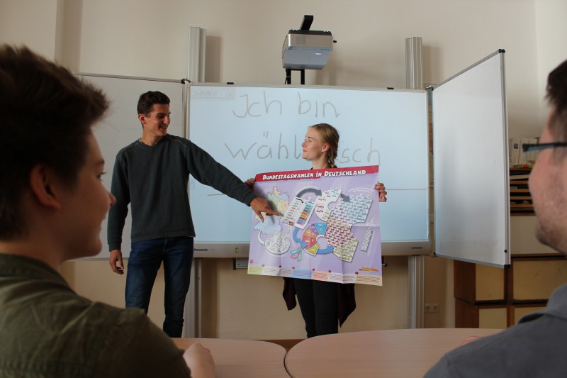 KI generiert: Das Bild zeigt eine Person, die vor einer Gruppe steht und eine Präsentation auf Deutsch hält. Auf der Tafel hinter ihr steht "Ich bin wählbar" und sie hält ein Plakat über Bundestagswahlen in Deutschland.