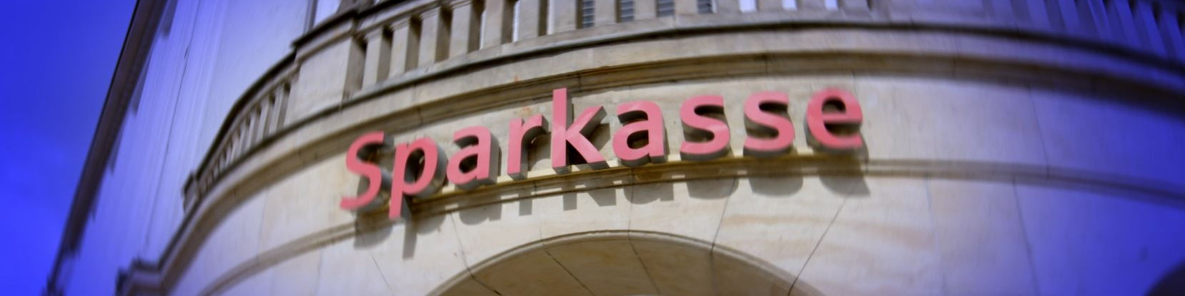 KI generiert: Das Bild zeigt die Fassade eines Gebäudes mit dem leuchtend roten Schriftzug "Sparkasse" darüber. Der Hauptinhalt des Bildes ist das Logo der Sparkasse, einer bekannten Bank in Deutschland.