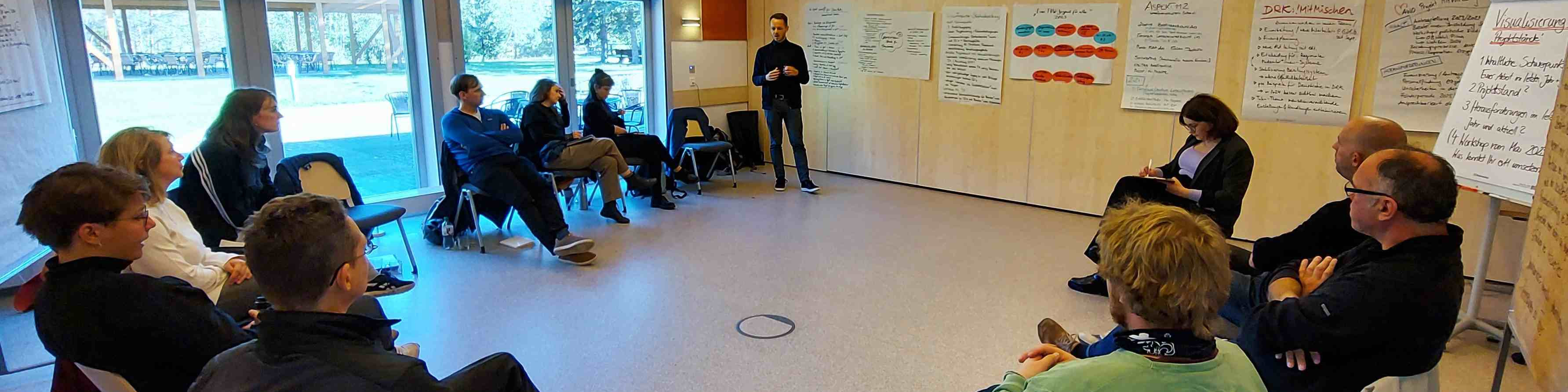 KI generiert: Das Bild zeigt eine Gruppe von Personen, die in einem hellen Raum mit großen Fenstern an einer Besprechung oder einem Workshop teilnehmen. Ein Mann steht in der Mitte und präsentiert etwas, während die anderen auf Stühlen sitzen und zuhören, wobei an den Wänden verschiedene Notizen und Diagramme an Whiteboards hängen.