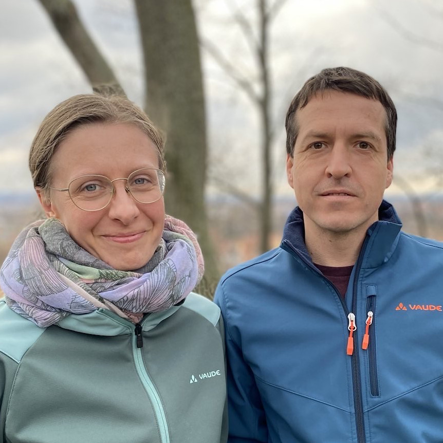 KI generiert: Das Bild zeigt zwei Personen, die im Freien stehen und in die Kamera lächeln. Beide tragen Outdoor-Jacken der Marke Vaude und stehen vor einem Hintergrund mit Bäumen und einer leicht bewölkten Landschaft.