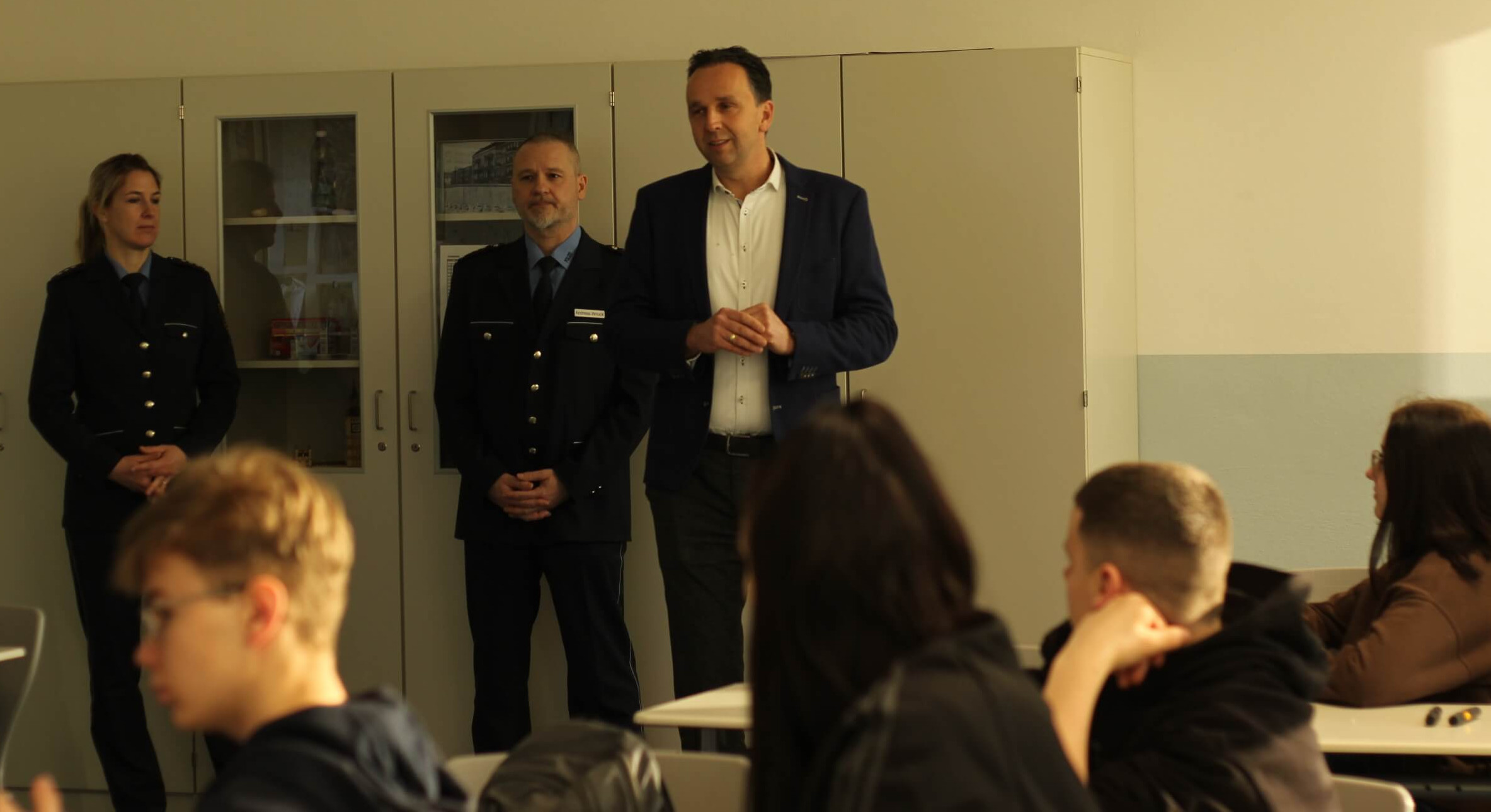 KI generiert: Das Bild zeigt eine Gruppe von Personen in einem Klassenzimmer, wobei drei Erwachsene vorne stehen und eine Gruppe von Schülern zuhört. Der Hauptinhalt des Bildes ist eine Unterrichts- oder Vortragssituation, bei der die Erwachsenen, darunter zwei in Uniform, mit den Schülern interagieren.