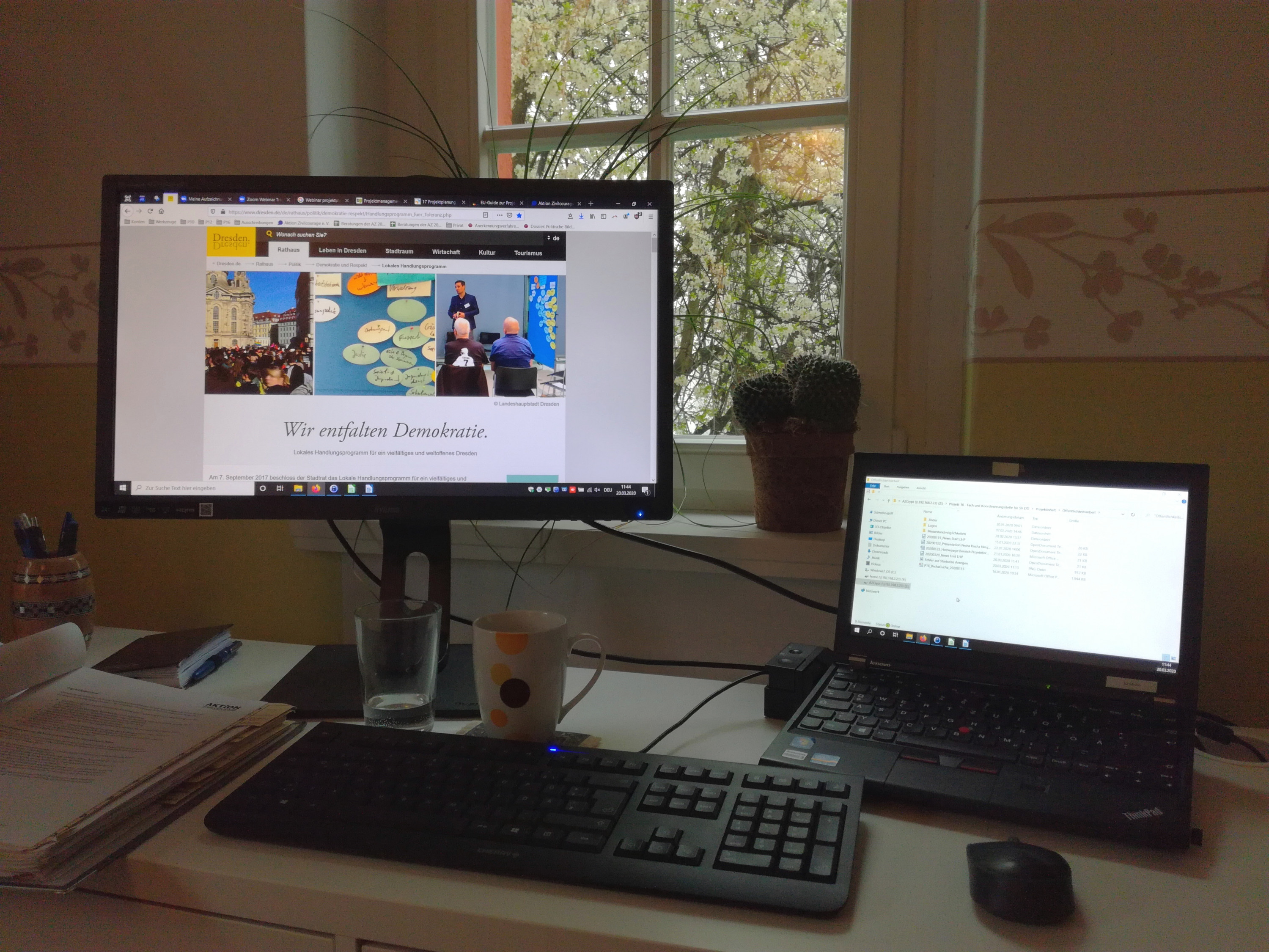 KI generiert: Das Bild zeigt einen Arbeitsplatz mit einem Desktop-Computer und einem Laptop auf einem Schreibtisch vor einem Fenster. Der Hauptinhalt des Bildes ist die Arbeitsumgebung mit den beiden Computern, wobei auf dem Bildschirm des Desktops eine Webseite mit dem Titel "Wir entfalten Demokratie" zu sehen ist.