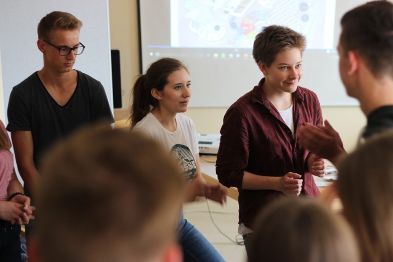 KI generiert: Das Bild zeigt eine Gruppe von Personen in einem Klassenzimmer, wobei einige im Vordergrund stehen und sich unterhalten, während andere im Hintergrund sitzen und zuhören. Der Hauptinhalt des Bildes ist eine interaktive Diskussion oder ein Workshop in einer Bildungs- oder Schulumgebung.