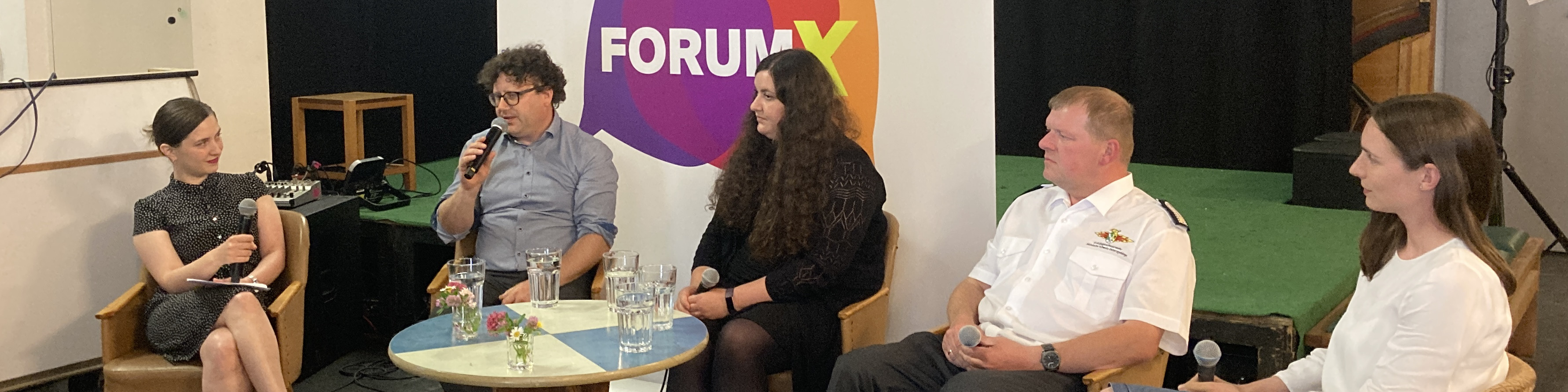 KI generiert: Das Bild zeigt eine Podiumsdiskussion mit fünf Personen, die auf der Bühne sitzen und Mikrofone verwenden. Im Hintergrund ist ein Banner mit der Aufschrift "FORUM X" zu sehen.