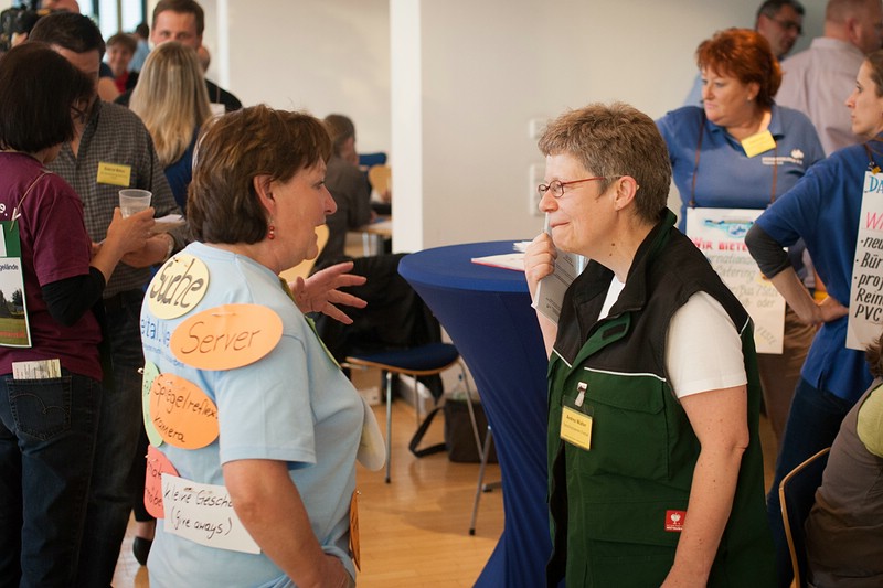 KI generiert: Das Bild zeigt eine Gruppe von Personen in einem Innenraum, die sich in kleinen Gruppen unterhalten. Im Vordergrund stehen zwei Frauen, eine mit einem T-Shirt mit Aufklebern und die andere mit einer Weste, die sich angeregt austauschen.