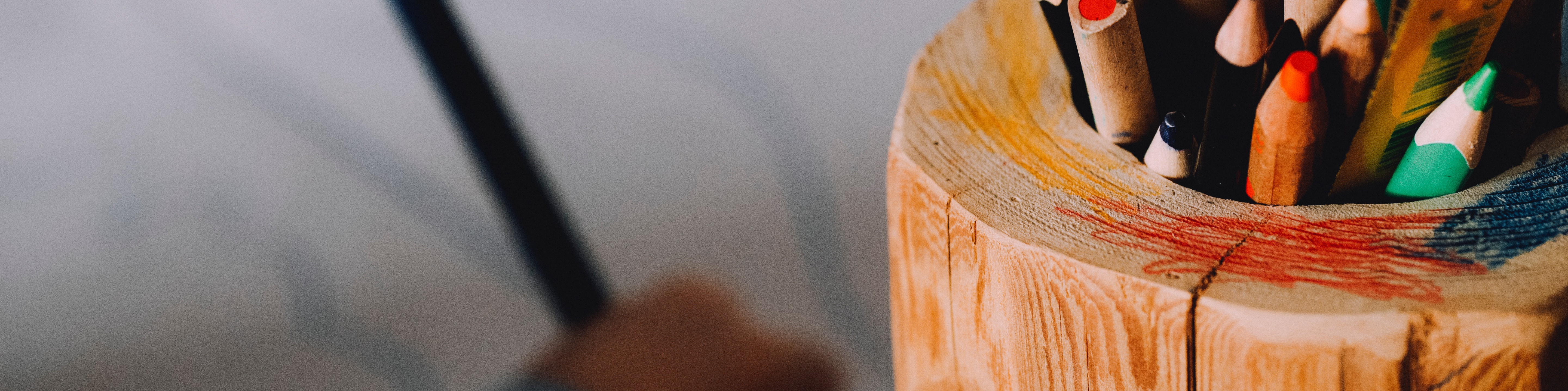 KI generiert: Das Bild zeigt einen Holzbecher, der als Stiftehalter dient und verschiedene bunte Stifte enthält. Im Hintergrund ist ein unscharfes Bild eines Stuhls zu erkennen.