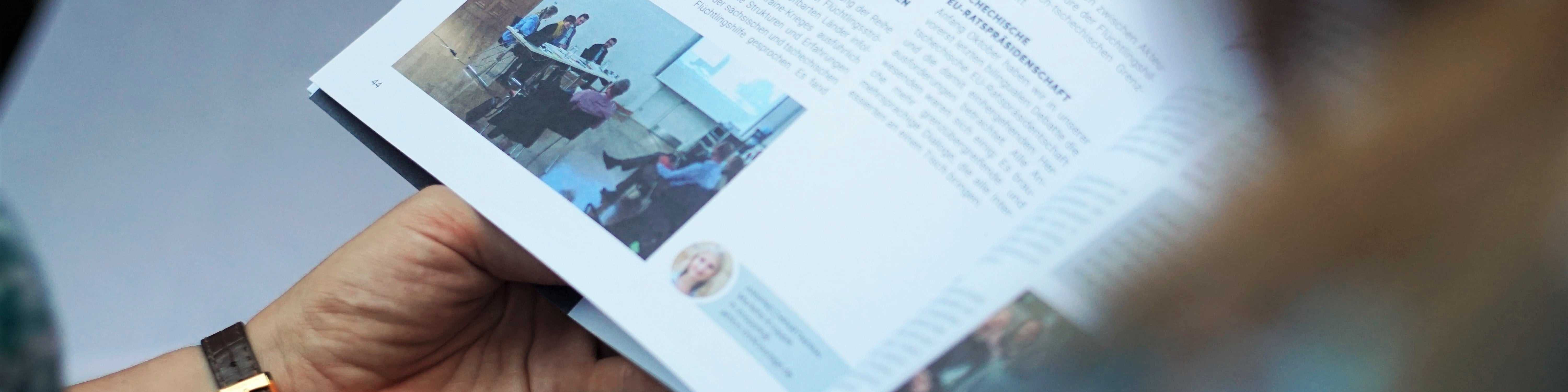 KI generiert: Das Bild zeigt eine Person, die ein Buch oder eine Broschüre mit einer Bildseite und Text hält. Der Hauptinhalt ist die Darstellung einer Seite mit einem Foto von Menschen, die an einem Tisch sitzen, und begleitendem Text.