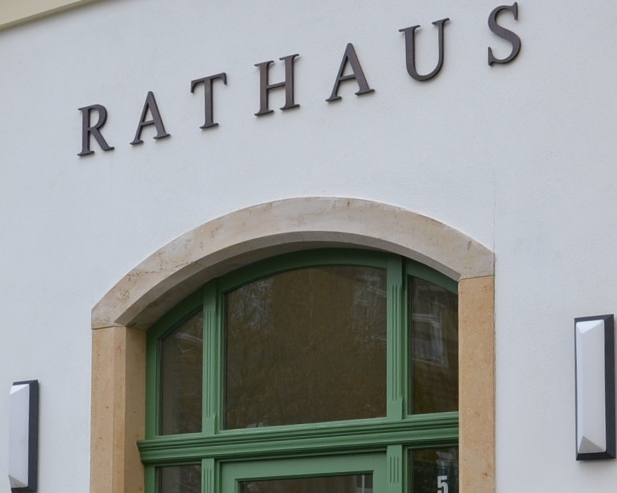 KI generiert: Das Bild zeigt die Fassade eines Gebäudes mit dem Namen "RATHAUS" in großen, dunklen Buchstaben an der Wand. Der Hauptinhalt des Bildes ist die Kennzeichnung des Gebäudes als Rathaus.