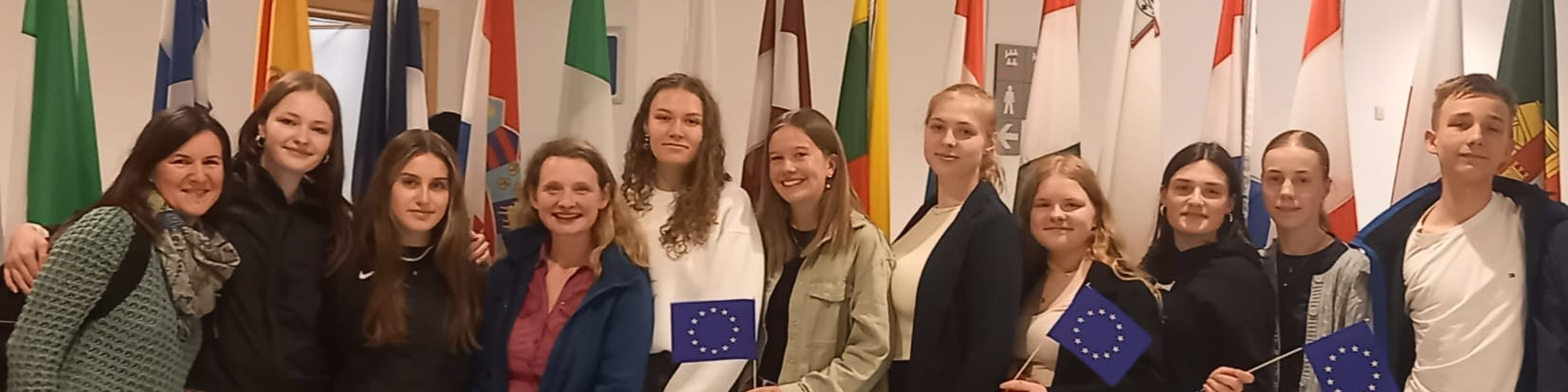 KI generiert: Das Bild zeigt eine Gruppe von Jugendlichen und einer erwachsenen Begleitperson, die vor einer Reihe von Flaggen verschiedener Länder posieren und einige von ihnen halten kleine EU-Flaggen in den Händen. Der Hauptinhalt des Bildes ist eine Gruppe von Menschen, die möglicherweise an einem internationalen oder europäischen Event teilnehmen.