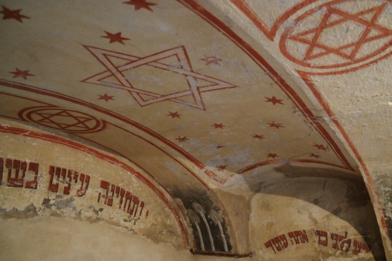 KI generiert: Das Bild zeigt eine alte, gewölbte Decke mit hebräischen Inschriften und roten Sternen von David. Der Hauptinhalt des Bildes sind die religiösen Symbole und Schriftzüge, die auf eine jüdische Tradition oder einen historischen Ort hinweisen.