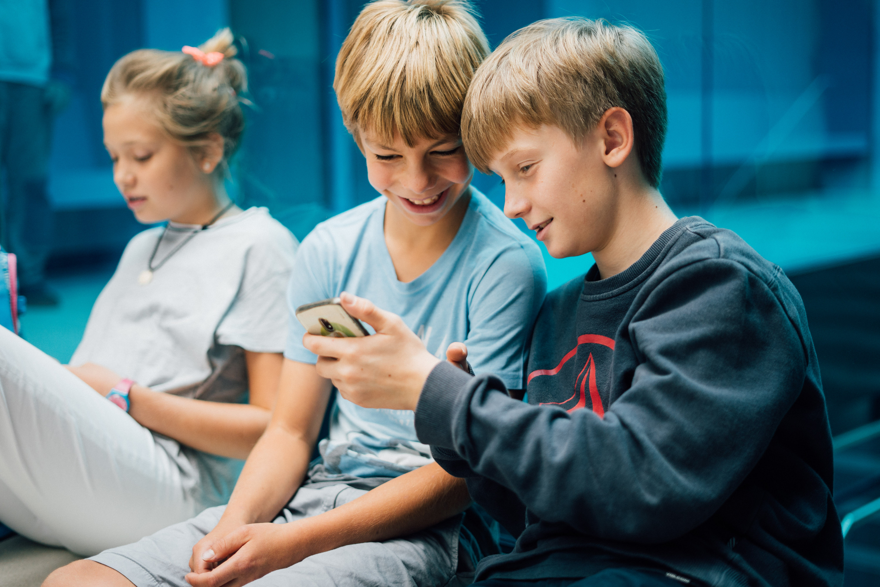 KI generiert: Das Bild zeigt drei Kinder, die eng beieinander sitzen und gemeinsam auf ein Smartphone schauen. Der Hauptinhalt des Bildes ist die Interaktion der Kinder mit dem Smartphone, was ihre Aufmerksamkeit und Neugier weckt.