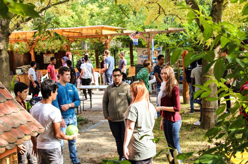 KI generiert: Das Bild zeigt eine Gruppe von Menschen, die sich in einem Wald oder Park bei einem Event oder einer Versammlung unterhalten. Im Hintergrund sind Holzhütten und ein Basketballkorb zu sehen.
