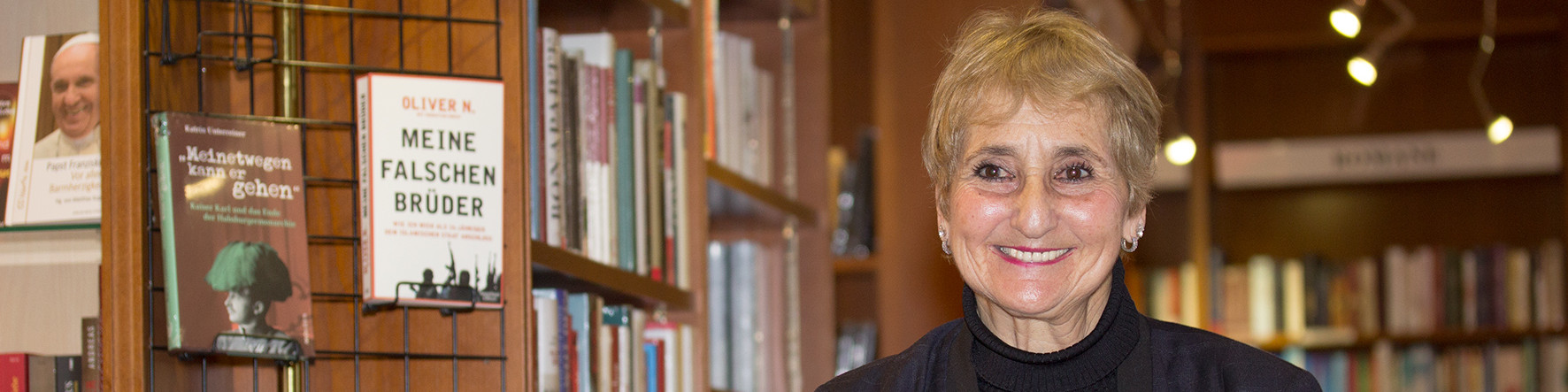 KI generiert: Das Bild zeigt eine lächelnde Frau in einem Buchladen oder einer Bibliothek, umgeben von Büchern und Regalen. Der Hauptinhalt des Bildes ist die Frau, die vor einem Regal mit verschiedenen Büchern steht.