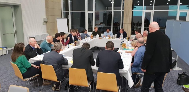 KI generiert: Das Bild zeigt eine Gruppe von Personen, die um einen langen, mit weißem Tuch bedeckten Tisch sitzen und sich in einer Besprechung oder einem Workshop befinden. Der Raum ist hell, mit großen Fenstern und Flipcharts an den Wänden.