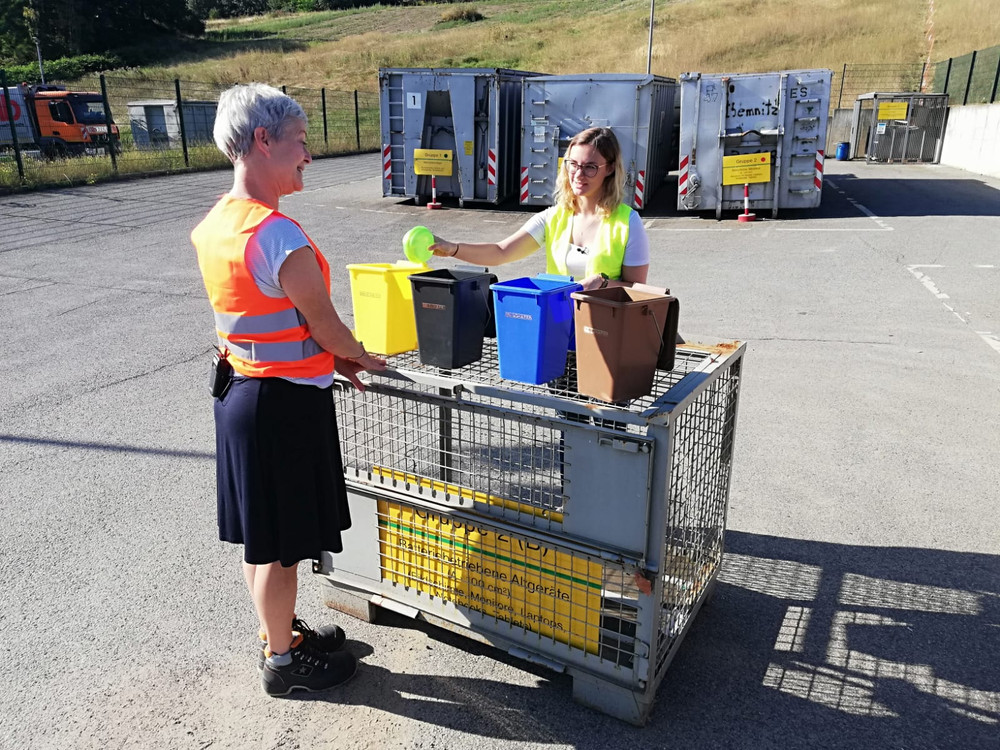 KI generiert: Das Bild zeigt zwei Personen in Warnwesten, die sich in einem Industrie- oder Logistikbereich unterhalten und dabei einen Rollcontainer mit verschiedenen Abfallbehältern und einem Pappkarton nutzen. Der Hauptinhalt des Bildes ist die Abfalltrennung und -entsorgung in einem gewerblichen Umfeld.