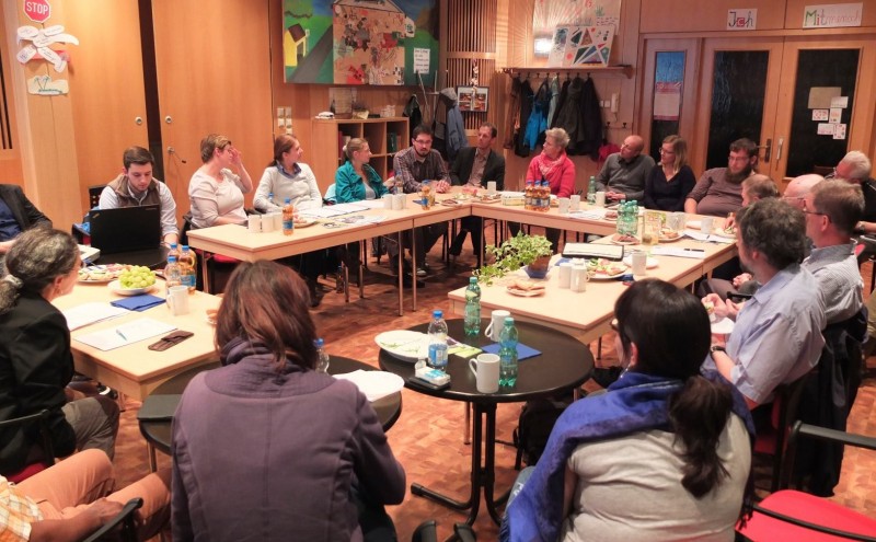 KI generiert: Das Bild zeigt eine Gruppe von Menschen, die in einem Raum mit Holzwänden und -möbeln an mehreren Tischen sitzen und sich unterhalten. Der Hauptinhalt des Bildes ist eine informelle Besprechung oder ein Treffen in einer gemütlichen, Gemeinschafts- oder Büroumgebung.